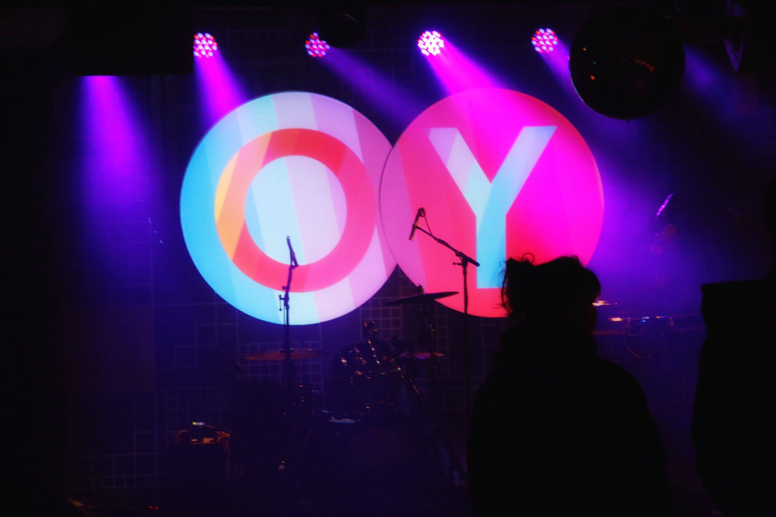 Foto von noch leerer Bühne vor dem OY-Konzert. Es sind zwei große, quasi schwebende Kreise mit den bunten Buchstaben OY zu sehen.